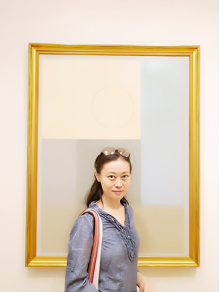 Photo of a smiling woman with long dark hair posed in front of an empty gold frame in an art gallery.